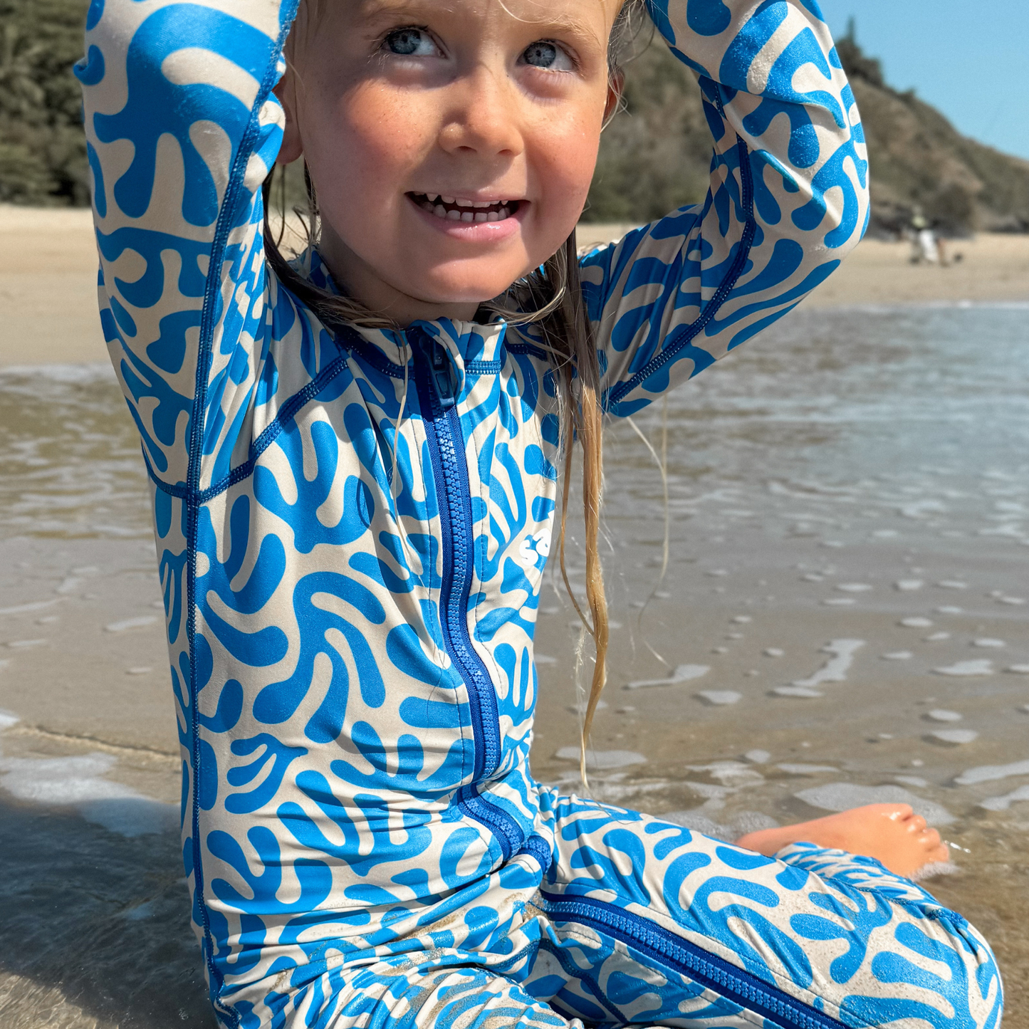 Child wearing a blue and white patterned stinger suit sitting on a beach.